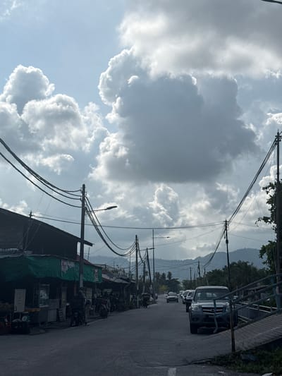 The image depicts a street scene in George Town, Malaysia. The street is lined with buildings and vehicles. The sky is cloudy and there are mountains in the distance. The street is populated with parked cars and motorbikes. Power lines run overhead. The image is shot from a low angle.