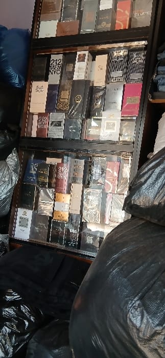 The image depicts an indoor scene in Sabon Gari South, Nigeria, featuring a multi-tiered, dark-framed display cabinet filled with numerous boxed perfumes or fragrances. The boxes are predominantly rectangular, many still sealed in clear cellophane, and are stacked closely together on at least four visible shelves within the cabinet. Identifiable labels include "212 NYC MEN," "Si," "WANTED," and "212 SEXY MEN." To the immediate left of the cabinet, a blue textile object is visible at the top, and crumpled black plastic bags are on the floor. To the right, a stack of folded dark garments, possibly jeans, is present on a shelf. A large, dark object, consistent with a bale or large bag wrapped in black plastic, occupies the right foreground, partially obscuring the lower right section of the display cabinet. Additional black plastic bags are visible on the floor in the lower left foreground. No individuals are present.