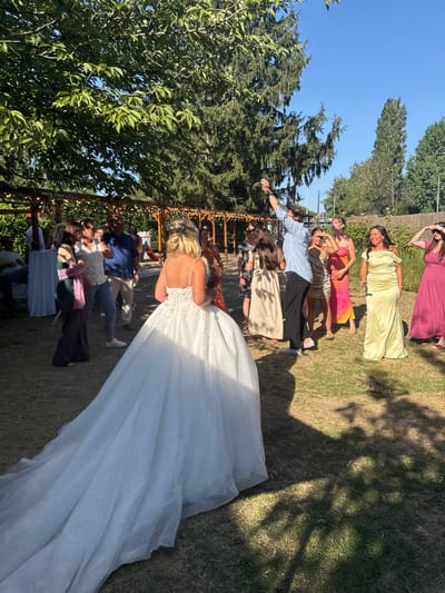 In Hem, France, a wedding reception is underway outdoors under a clear blue sky. A bride in a white gown stands with her back to the camera, her train spread across the lawn. Guests mingle in the background, some in formal attire, others more casually dressed. A man in a light blue shirt and dark pants is seen with his arm raised. The scene is set in a grassy area with trees and a wooden pergola adorned with string lights.