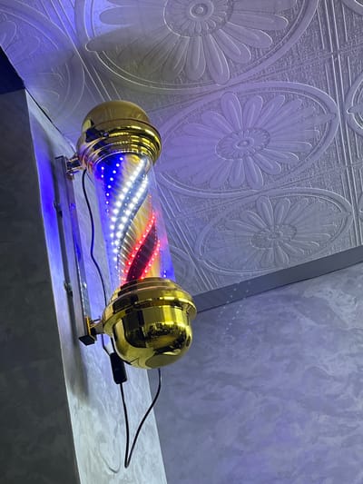A barber pole with red, white, and blue lights is mounted on a wall in what appears to be a barbershop. The pole is cylindrical and encased in a clear tube with rotating lights. It has golden finials at the top and bottom. The wall behind the pole has textured wallpaper with a floral pattern. The floor is a light-colored, textured surface. The lighting in the scene is predominantly from the barber pole and ambient blue and purple light reflecting off the walls and ceiling. The image is taken from a low angle, looking up at the barber pole. There are no people visible in the image. The specific location in Al Machouar – Stinia, Morocco is not discernible from the image itself.