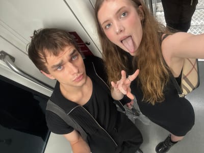 In the city of Xuhui District, China, two individuals, a man and a woman, are captured in a close-up selfie taken from a high angle, likely inside a mode of public transportation, suggested by the metallic handrail and patterned flooring visible. The man, positioned on the left, has short brown hair and striking blue eyes. He is wearing a black t-shirt layered with a black zipped vest. The woman, to the right, has long, light brown hair and is sticking her tongue out towards the camera, with a ring on her left index finger. She is wearing a black dress and a distinctive checkered shoulder bag. The background appears to be the interior of a train or subway car, indicated by the door panel with red signage and a metal handrail.