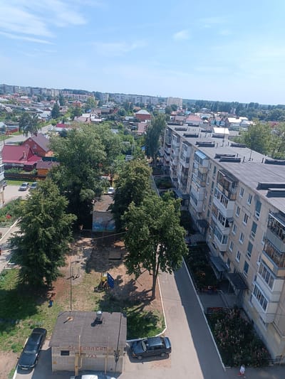 This is an aerial, wide-angle, eye-level, daytime photograph taken in Dimitrovgrad, Russia, showing a residential area. The foreground features a multi-story apartment building on the right, characterized by its yellow facade, numerous balconies, and dark grey flat roof. Adjacent to the building is a paved walkway lined with flower beds. In the middle ground, a playground with yellow exercise equipment and a blue structure is visible, surrounded by sandy ground and green trees. A small brick building with a dark roof, identified as a gas utility building with Russian text reading "ГАЗ" (GAZ), is situated to the left of the playground. Two dark-colored sedans are parked near this building. Further back, a mixture of smaller residential houses with red and white roofs and more trees populate the landscape. The background displays a distant view of urban structures and a clear blue sky with sparse clouds.