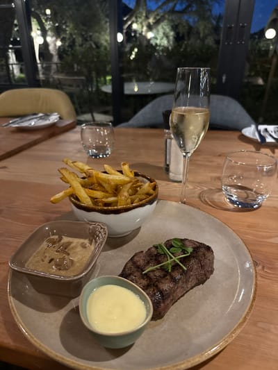The image shows a table setting at a restaurant, likely indoors with a view of an outdoor area at night. The table is made of wood. On the table, there's a plate containing a piece of grilled steak garnished with a small green sprig, a small bowl of yellow sauce, and a small dish of mushroom sauce. Next to the plate is a white bowl filled with french fries. A flute glass containing a light-colored beverage and an empty glass tumbler are also present. The restaurant appears to be located in Paarl, South Africa, judging by the general aesthetic of the setting. There are no people visible in the image. The lighting is dim, suggesting an evening meal.