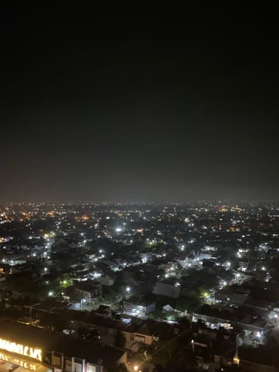 This is an elevated nighttime photograph depicting a sprawling residential urban area in the city of Entalsewu, Indonesia. The scene consists of a densely packed network of illuminated buildings, primarily houses and low-rise structures, extending towards the horizon under a dark, hazy sky. Numerous artificial light sources, predominantly white and yellow, punctuate the darkness, indicating individual residences, street illumination, and some larger establishments. Dark, irregular shapes consistent with trees or vegetation are interspersed among the structures. A faint, thin white streak is present in the upper-left sky quadrant. In the bottom foreground, elements of a larger structure are partially visible, featuring bright yellow illumination suggestive of signage or interior lighting. No individuals or specific human actions are identifiable within the frame.