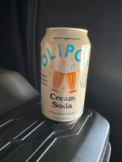 The media file shows a can of Olipop Cream Soda resting on a dark, textured surface, possibly the interior of a vehicle. The can is the primary subject, oriented horizontally. The lighting suggests it is daytime, though no specific time can be determined. The can's label is partially visible, featuring the brand name "OLIPOP" in large blue lettering. Below this, it indicates "Cream Soda" and "A New Kind of Soda." Smaller text mentions "Supports Digestive Health" and lists ingredients like "Probiotics," "Botanicals," and "Fiber." There are also illustrations of stylized glasses with orange accents. The background is dark and out of focus, providing minimal contextual information about the immediate surroundings, other than the textured surface on which the can rests. There are no people or animals visible. The overall impression is a simple product shot with a focus on the beverage can.