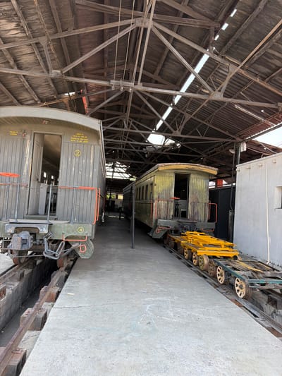 The image shows a collection of old railroad cars housed inside a large, weathered, open-sided building with a metal frame and corrugated metal roof. The roof has some holes allowing daylight to filter through. The railroad cars are of different designs and states of repair. One is painted grey and bears the text "REP-G CUA-10-08-M PESO 16 900 KGS LGO 11 89 MTS". Another is painted yellow. The location is likely a museum or historical site in Cuautla, Mexico, referenced by the “CUA” on the railroad car. A concrete walkway runs alongside the railroad cars.