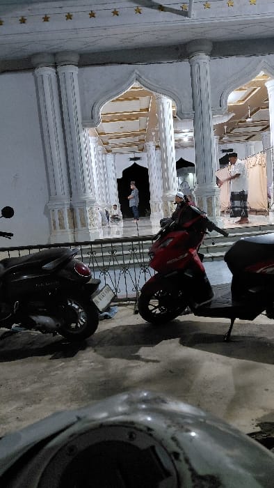 The scene depicts the exterior of a mosque or religious building at night in Keudealue Ie Puteh, Indonesia, viewed from a low vantage point. In the immediate foreground, the top-front section of a motorcycle, light-colored and out of focus, is visible. The ground is dark concrete.
Parked on the concrete in the midground are two scooters. To the left, a dark-colored scooter, possibly a Honda Vario, is partially visible, facing left. Its license plate, "BL 6163 KHD," is legible. To the right, a red scooter, also possibly a Honda Vario, is parked facing right, resting on its side stand. A black metal fence or railing with decorative elements separates the parking area from the building's entrance.
The building itself features white fluted columns and multiple ornate pointed arches forming entrances. The interior is brightly lit, revealing a white ceiling with geometric patterns and golden or yellowish decorative stars on overhead beams.
Several individuals are present. Inside the building, from left to right: a child in a light shirt and dark pants stands near an archway; an adult male in a light-colored top and dark lower garment, wearing a cap, stands further inside; another adult male wearing a white shirt and a checkered sarong is standing, holding a brown rectangular object, possibly a book or folder. Closer to the foreground, near the red scooter and behind the fence, an adult male wearing a white kufi cap and a dark top is partially visible. Further indistinct figures are discernible within the illuminated interior of the building.