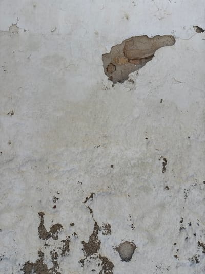 This image shows a close-up of a weathered, textured wall. The wall is predominantly white with areas of damage and discoloration. There are several large patches where the white plaster has chipped away, revealing the rough, grey, and brown material underneath. Some of these exposed areas are deep, showing the layering of the underlying structure, while others are shallow pits. The surface of the white plaster is uneven, with fine cracks visible across its entirety, suggesting age and exposure to the elements. There are also small, scattered dark spots, possibly from dirt or mildew. The lighting appears natural and diffused, suggesting it was taken during the daytime, with no strong shadows indicating a specific time. The texture of the wall suggests it is an exterior surface, possibly of a building, given the wear and tear. There are no people, objects, or discernible text in the image. The overall impression is one of decay and the passage of time on a man-made surface. The context of Vilanova i la Geltrú, Spain, implies this could be part of an older building in a coastal or Mediterranean environment, which often contributes to such weathering patterns.