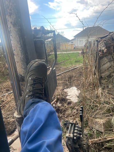 The image shows a close-up view from inside a piece of heavy machinery, possibly a digger or tractor. The viewer is positioned within the cab, looking out. In the foreground, a person's foot, clad in a work boot and blue pants, rests on a metal pedal or footrest.
Outside the machinery, the environment appears to be a rural or suburban setting. There is a patch of grassy land, some tangled dry vegetation, and a building in the background. The building is light-colored and has a gabled roof. Power lines stretch across the upper left of the frame, against a sky with blue and white clouds.
The location is Поедуги, Russia.