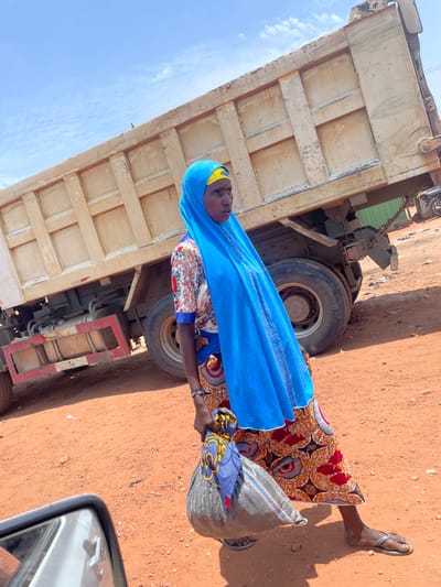 A woman in Kwali, Nigeria, stands beside a large truck. She wears a brightly patterned dress and a blue headscarf. She carries a gray bag. The ground is reddish dirt. A portion of a car is visible in the lower right corner of the image.
