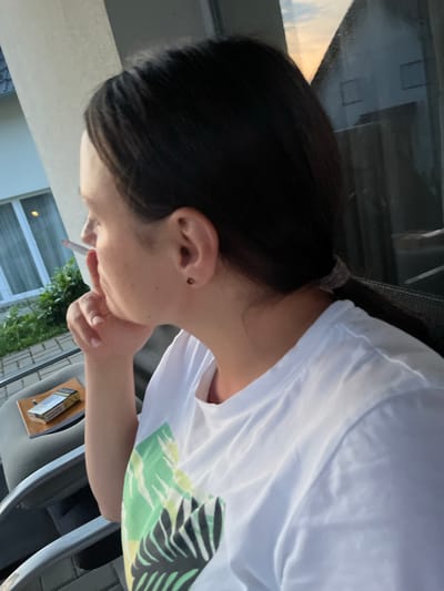 A woman is viewed from the side, smoking a cigarette and looking out a window. She has dark hair pulled back in a ponytail and is wearing a white t-shirt with a green and black leaf pattern. She has a small black stud earring in her right ear. A pack of cigarettes and a small book or notebook are on a tray to her left. Outside the window, there is greenery and a building. The setting appears to be outdoors, possibly on a balcony or patio. The woman is in Milná, Czechia.