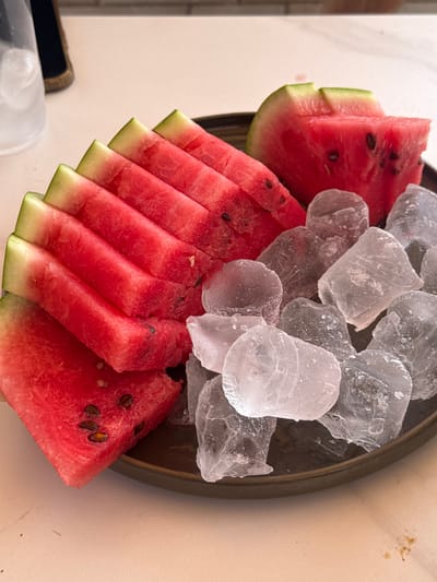 This close-up image showcases a serving of fresh watermelon slices accompanied by ice cubes on a dark plate. The watermelon is cut into triangular wedges and arranged in a fan-like pattern. The vibrant red flesh of the fruit is visible, along with scattered dark seeds. The rind shows the characteristic green and white layers. The ice cubes are clear and irregular in shape, piled on one side of the plate, suggesting a refreshing treat. The setting appears to be an indoor surface, possibly a table, with a neutral, light-colored background that has some subtle textures and marks. The lighting suggests daylight, possibly a warm afternoon. No people or specific location cues are visible beyond what can be inferred about the food and its presentation. The image captures a simple, appealing visual of a chilled fruit snack.