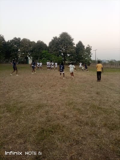 This image depicts an outdoor sports activity taking place on a dry, open field with sparse grass. Approximately 15-20 individuals are visible, engaged in or observing a game, likely soccer or rugby.

In the mid-ground, a person wearing a dark jersey with the number "3" on the back and white shorts is seen, facing away from the camera and seemingly participating in the play. Close by, another player in a white jersey with the number "7" and white shorts is in motion, appearing to run with or towards a light-colored ball. Several other players in dark and white attire are scattered across the field, actively involved in the game.

To the left-center in the background, a group of about 10-12 people, mostly dressed in white shirts and shorts, are gathered, possibly forming a huddle or observing the ongoing activity. On the far right, a person in a yellow t-shirt and dark trousers stands facing away, watching the event.

The background features a dense line of trees, suggesting a natural or semi-rural environment. Beyond the trees on the right, some distant structures and a concrete wall are visible, along with a utility pole. The sky is bright and clear, indicating fair weather, and the soft lighting suggests it is either late afternoon or early evening. The ground is predominantly brown and parched, with patches of greener vegetation. The overall scene portrays an active community sports event. The visible text "Infinix NOTE 8i" appears in the bottom left corner, identifying the camera used. The activity is set in Ayere, Nigeria.