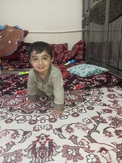 A young child, likely a toddler, is crawling on a patterned rug in an interior setting. The child is wearing a striped pajama set and is smiling. The rug has a repeating pattern of floral and abstract designs in beige and red tones. Behind the child is a bed or seating area covered with cushions and blankets. There is a brown blanket, and red cushions with floral patterns. The cushions appear to be on a floral pattern mat, where a blue pillow with white flowers also lies. In the background, a dark wooden cabinet or wardrobe with ornate detailing is visible against a plain white wall. Based on the presence of a rug and interior furnishings, this scene takes place inside a home or dwelling. There is no specific information to confirm the Galaosiyo, Uzbekistan location.