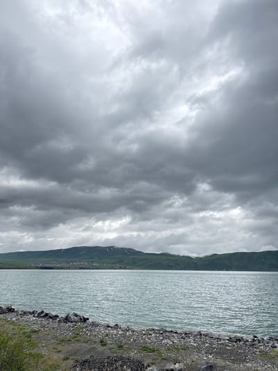 The image shows a body of water under a cloudy sky, potentially located in the vicinity of Tsamakaberd, Armenia. A mountain range is visible on the opposite side of the water. The shoreline consists of a rocky terrain with some vegetation.