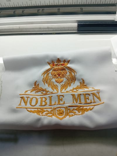 A white cloth embroidered with a golden emblem is laid out on a white surface. The emblem features a lion's head wearing a crown, with ornate gold detailing surrounding it, including floral motifs and a shield with a crown. The words "NOBLE MEN" are embroidered in gold beneath the lion's head. The cloth appears to be part of a larger piece, with a raw edge visible on the left side. In the background, a metallic ruler with markings is visible, suggesting a crafting or measuring context. The overall scene is clean and well-lit.