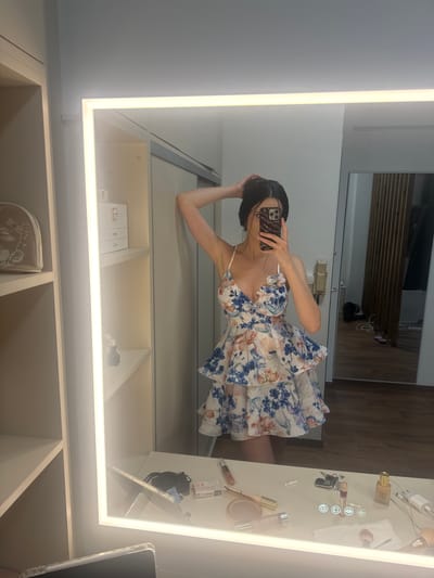 A woman stands in a brightly lit room, likely a dressing area, as reflected in a large, illuminated mirror. She is wearing a white floral print dress with ruffles and thin straps. Her hair is dark and pulled back, and she is holding her phone to take a selfie. To her left, there are shelves with various items, including a makeup bag and a jewelry box. The mirror reflects a vanity table with makeup, keys, and other personal items. The room appears modern with wooden flooring and a light-colored wall. The scene is located in Agios Dimitrios, Greece.