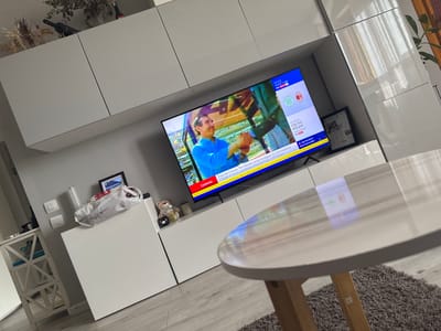 The image captures a modern living room interior, likely in an apartment. The scene is dominated by a large flat-screen television displaying a sports broadcast, possibly cycling given the text "Ciclismo" and information about the Tour de France. The television is mounted above a white media console. To the left of the console is a tall, white shelf unit with geometric patterns, holding various items. On top of the media console, there are a few decorative objects including a teddy bear, a glass jar with gold-colored contents, and a small black figurine. A white plastic bag and a framed picture are also visible on the console. To the right of the television, a section of glossy white cabinetry is present, reflecting light. In the foreground, a round, light-colored coffee table with wooden legs sits on a grey rug. The overall impression is one of a clean, contemporary living space. There is no information in the image that would allow for definitive identification of the location as Ferrara, Italy.
