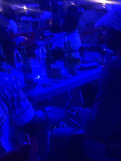 The image shows a dimly lit, blurry scene with strong blue lighting. In the foreground, a person is partially visible wearing a blue garment with what appears to be the word "MARINI" and stars on it, suggesting military or patriotic attire. The person is facing towards the right of the frame. Behind them, a table is laden with bottles, possibly drinks, and some items that are indistinct due to the blur. Other figures are present in the background, also indistinct and blurred, indicating a crowded environment. The overall atmosphere suggests a social gathering or event, possibly a bar or club, in Abuja, Nigeria, given the blue lighting and the presence of drinks and people. The motion blur suggests movement within the scene, possibly during a performance or active conversation.