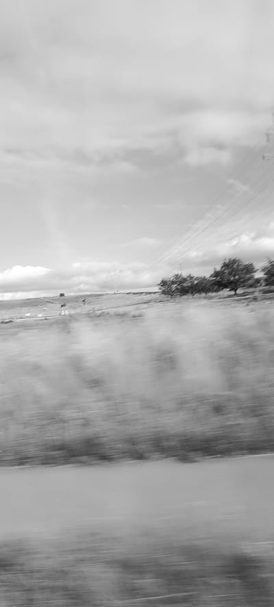The image captures a motion-blurred view of a rural landscape. The scene is dominated by a sky filled with clouds, with a faint rainbow visible on the left side. Power lines stretch diagonally across the frame from the upper right, converging towards the left horizon. Below the sky, a grassy field is depicted with trees scattered throughout. In the foreground, a road or path is visible, also blurred due to motion. The image appears to have been taken from a moving vehicle, likely a car, traveling along a road in the outskirts of Schwaikheim, Germany, given the presence of agricultural land and infrastructure typical of such areas. There are no discernible people or specific actions taking place.