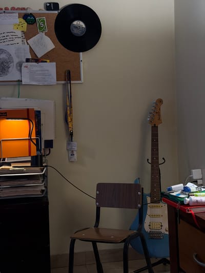 The image shows a cluttered room with a guitar mounted on the wall, alongside a vinyl record attached to a bulletin board. Below the record, there are papers, cards, and notes pinned on the board. To the right, a lanyard with a badge hangs above a stack of notebooks and folders. A chair with a wooden backrest is situated next to a desk where toiletries are placed. The overall lighting in the room appears dim.