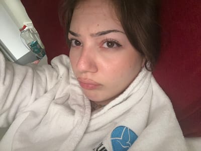 A fair-skinned woman lies on a red surface, possibly a couch or bed, wearing a white bathrobe with a blue logo. She has brown hair and defined eyebrows, and her eyes are brown with subtle eyeliner. Her lips are full, and she appears to be looking directly at the camera with a relaxed expression. In the background, a small glimpse of what seems to be a side table with bottles and a white surface can be seen. The photo is taken from a slightly angled perspective, with the woman's head tilted.