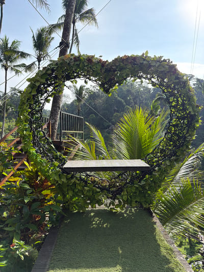 This image captures an outdoor scenic overlook in Tegal Suci, Indonesia, featuring a large, heart-shaped decorative archway. The archway, constructed from a dark, intricate woven frame, is densely covered with green and some reddish-brown artificial foliage. Integrated into the lower curve of the heart is a horizontally planked platform of light-colored composite material with a distinct circular pattern, intended for standing. This structure rests on a ground-level platform, partially covered with artificial green turf and bordered by wooden planks.
To the left, a wooden staircase with dark metal railings ascends towards an elevated wooden platform. A tall, slender tree trunk is visible near the staircase, secured by multiple tensioned cables extending upwards out of frame. The immediate environment is characterized by lush tropical vegetation, including broad-leaved plants displaying green, red, and orange hues. Directly behind the heart structure, the large, vibrant green fronds of a palm tree are prominently illuminated by sunlight. The background reveals a dense tropical forest covering a sloping landscape, dotted with numerous additional palm trees. The sky above is clear and bright blue, indicating fair weather. Strong sunlight illuminates the scene from the upper right, casting partial shadows on the artificial turf. No individuals are present in the image.