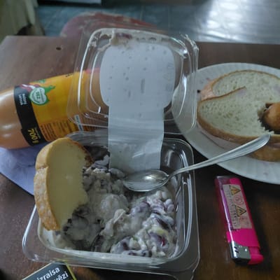 A close-up, overhead shot shows food items and a lighter on a wooden surface, likely a table. In the foreground, a clear plastic container holds a portion of potato salad with red beans and a piece of bread is resting on the edge. To the right, a pink disposable lighter with warning labels is placed next to a plate of sliced bread topped with what appears to be meat. Behind the container, a partially visible sausage with a yellow wrapper is lying on a blue paper towel. The background is blurred, showing a light-colored surface. The image is taken in Skuķīši, Latvia.