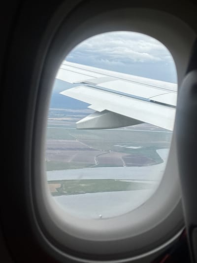 The view from an airplane window reveals a wing and a landscape below. The landscape consists of fields and a river. It's difficult to determine the exact location, the image does not show the location of Montijo, Portugal. No people, subjects, objects, actions, or interactions are visible in the image. The environment is the interior of the airplane and the landscape seen through the window.