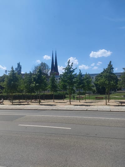 The image shows a street in Chemnitz, Germany, with a park area on the right side and a building with two spires in the background. The park has several trees, benches, and a playground. There is one person sitting on a bench in the park. The street in the foreground is paved and has a white line marking a lane. The sky is blue with a few white clouds.