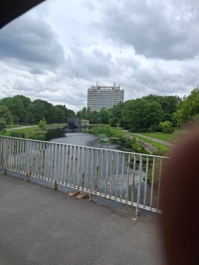 The image depicts a view from a bridge overlooking a canal or river in the city of Piła, Poland. In the foreground, a grey concrete walkway leads to a rusty metal railing that extends across the bottom of the frame. Beyond the railing, the dark water of the canal is visible, dotted with patches of green aquatic plants. The canal banks are lined with lush green grass and trees. In the middle ground, a curved, dark structure, possibly a part of a dam or water control mechanism, sits on the canal. Across the canal, a path winds through a park area with benches and lampposts. In the background, a tall, light-colored building with many windows stands prominently, its facade reflecting the overcast sky. Trees and foliage surround the building and extend to the edges of the frame. The sky is heavily clouded with grey and white masses. A dark, out-of-focus object, possibly a finger or hand, intrudes into the right side of the foreground. The scene appears to be captured during daylight under overcast conditions, suggesting a temperate climate.