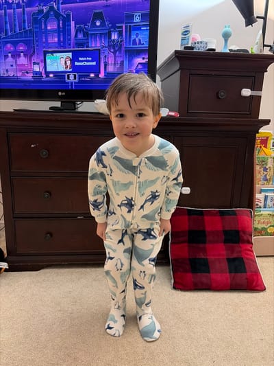 A young child, approximately toddler-aged, with light skin and dark hair, stands centrally on a light beige carpet, smiling directly at the camera. The child is dressed in a full-body pajama suit featuring a white background patterned with blue marine animals, including whales, orcas, and penguins, with integrated footies. Behind the child is a dark brown wooden dresser with four visible drawers, each equipped with a dark circular knob. A white child safety latch is affixed to the upper right dresser drawer. A black LG flat-screen television rests on the dresser, displaying an animated city scene with a purple sky, overlaid with an interface indicating "Watch free RokuChannel" and a small thumbnail image of two individuals. To the right of the dresser, a partial view of a light-colored bookshelf stocked with children's books is present. On the dresser's top surface, to the right of the television, various items are visible, including a white tube labeled "Cetaphil." On the carpet to the child's right is a red and black buffalo plaid cushion with white trim. The specific location within Woodstock, Canada, is not discernable from visual information in the image.
