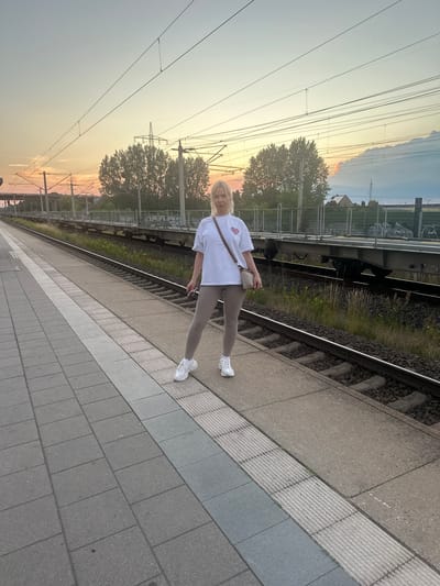 In Wustermark, Germany, a woman stands on a train platform with a sunset in the background. She is wearing a white t-shirt, gray leggings, and white sneakers. She has a small, quilted handbag slung across her chest. The platform is made of concrete tiles, with textured safety strips along the edge. Train tracks run alongside the platform, with a train on the other side of the tracks. Overhead, power lines stretch across the sky, supported by pylons. The sky displays a gradient of colors from pale blue to orange and pink, indicating the sun is setting.