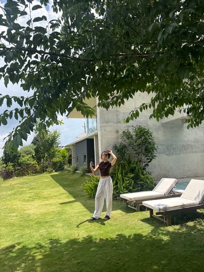A young woman stands on a grassy lawn in front of a modern concrete building, holding a smartphone to take a selfie. She is wearing a dark brown crop top and white patterned pants. Two lounge chairs with white cushions are situated on the grass next to a swimming pool. Lush green trees and bushes surround the area, with a large tree branch extending into the foreground, casting shadows on the lawn. The building has a minimalist design with concrete walls and large windows, and appears to be located in a suburban or rural setting, possibly in Đa Lộc, Vietnam, given the vegetation and architectural style. The sky is partly cloudy.