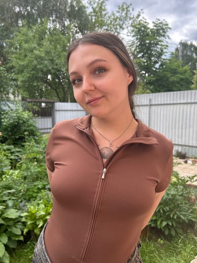 A young woman with light brown hair and green eyes is captured in a close-up, sideways shot. She is wearing a brown zip-up top and a patterned skirt. A delicate necklace with a small heart pendant rests on her collarbone, and a tattoo is visible on her chest. Her expression is relaxed and she appears to be smiling faintly.
The background suggests an outdoor setting, possibly a garden or backyard, with lush green foliage and trees. A white fence or building is visible behind her, and the sky is overcast, indicating a cloudy day. The overall mood is casual and serene. The image was taken in Chashnikovo, Russia. There is no visible text in the image.