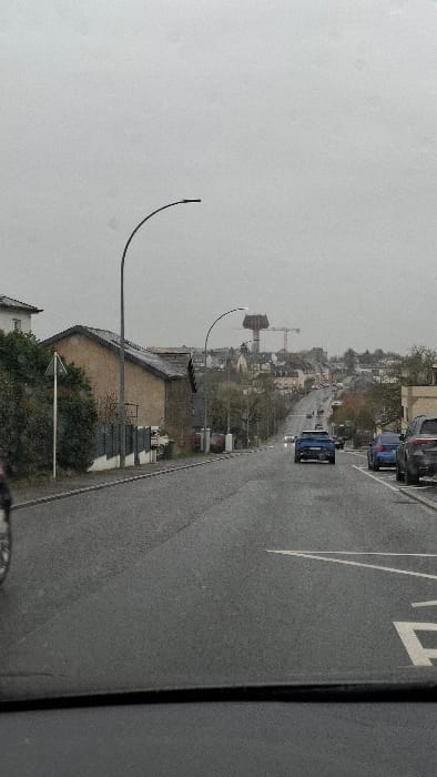 This image depicts a street scene in Frisange, Luxembourg, on an overcast day with a wet asphalt road, indicating recent or ongoing precipitation. The perspective is from inside a vehicle, looking uphill.
In the immediate foreground, a dark horizontal line consistent with a vehicle's dashboard is visible at the bottom. To the left, a portion of a bicycle's front wheel and fork is partially visible. On the right side of the road, white "P" (parking) road markings are present on the asphalt, next to a curb.
Along the left side of the street, a light brown residential building with a dark grey gabled roof is prominent, fronted by a dark fence and dense green shrubs. A white triangular street sign on a pole is positioned in front of the shrubs. A tall, black, curved street light pole stands near the center-left.
On the right side of the street, multiple vehicles are parked parallel to the curb, including a blue sedan and a dark grey SUV. A light-colored building is visible behind the parked cars. Another identical curved street light pole is positioned further uphill on the right.
The two-lane asphalt road ascends steadily towards the horizon. A blue SUV is visible in the middle of the road, driving uphill, with its taillights illuminated. Other vehicles are visible further up the incline, some displaying headlights, indicating traffic moving in both directions.
In the distant background, atop a hill, a distinctive, dark, "mushroom-shaped" or "tree-like" water tower stands prominently. Two yellow construction cranes are visible near the water tower, alongside a cluster of residential and commercial buildings forming a small townscape under the uniformly grey, overcast sky.