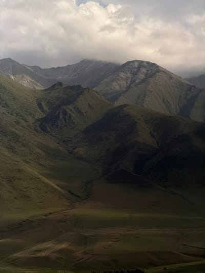 This is a still, vertically oriented, wide-angle photograph of a mountainous landscape. The scene is dominated by steep, green slopes of grass and rock, with distinct ridges and valleys carved into them. The lighting suggests it is daytime, with clouds obscuring parts of the sky and casting shadows on the mountainside. The overall mood is one of natural grandeur and ruggedness. The image is a landscape scene, with no people or discernible objects other than the natural topography. The location is Karagai Bulak, Kyrgyzstan, a region known for its dramatic mountain terrain. The weather appears to be mixed, with sunshine breaking through cloudy skies. There is no visible text in the image.