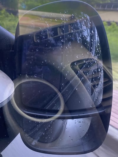 The image is a close-up, angled shot of a car's side mirror, reflecting the interior dashboard. The mirror's surface is covered with numerous water droplets, suggesting it is raining or has recently rained. The reflection shows part of the car's dashboard, including air vents and a circular vent or dial. The exterior environment reflected in the upper portion of the mirror appears to be a green, leafy area with some structures in the background, possibly a road or walkway. The overall impression is that of a vehicle parked outdoors in Pukhivka, Ukraine, during inclement weather.