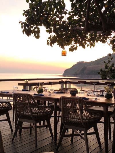 This image depicts an outdoor dining scene set on a wooden deck overlooking a calm body of water. Several wooden tables and chairs are arranged, each set with place settings including glasses, napkins, and cutlery. A small vase with greenery sits on one of the tables. Above, tree branches with lush green leaves hang over the scene, with a decorative lantern suspended from one of the branches.
The backdrop is a serene coastal landscape with a gently sloping hill covered in vegetation. The sun appears to be setting or rising, casting a warm, soft glow across the sky and water, with hues of orange and pink dominating the horizon. The atmosphere suggests a tranquil and inviting setting, likely a restaurant or resort, ready for diners to enjoy the scenic view. The location is identified as Labuan Bajo, Indonesia. No people are visible in the frame. A small sign on one of the tables reads "Ibu Sika". The weather appears clear and calm.
