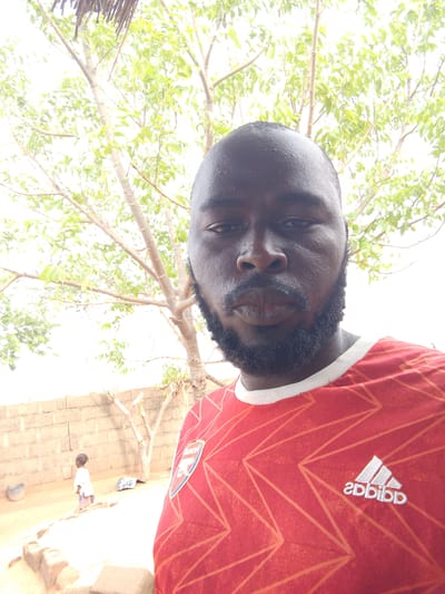 A man with a beard and bald head is in the foreground, wearing a red Arsenal soccer jersey. In the background is a tree with light green leaves and a wall made of beige brick. To the left of the wall is a small child wearing a white top. The scene appears to be outside in daylight. Given the prompt, this is presumably in Mulke Barikin Sayejo, Nigeria.