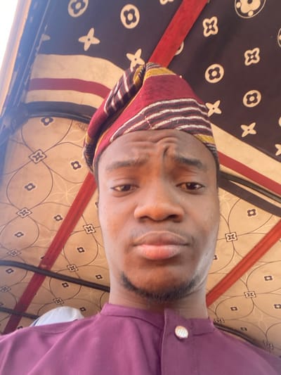 A dark-complexioned male, appearing as a young adult, is captured in a close-up, front-facing shot. He wears a maroon and yellow patterned traditional Nigerian cap (fila) and a maroon-purple traditional garment featuring a band collar and a single visible circular metallic button on the placket. The subject maintains direct eye contact with the camera, displaying a neutral facial expression with subtly pursed lips and a slight furrow in the forehead. Light stubble is present on his chin and jawline. The background consists of the interior canopy of a vehicle, characterized by a dark fabric adorned with repetitive light-colored cloverleaf and geometric patterns, white wavy stripes, and red fabric straps serving as structural or decorative elements. This setting is consistent with the passenger compartment of a commercial tricycle (keke napep), a prevalent mode of transport in Nigeria. The location is within Nigeria.