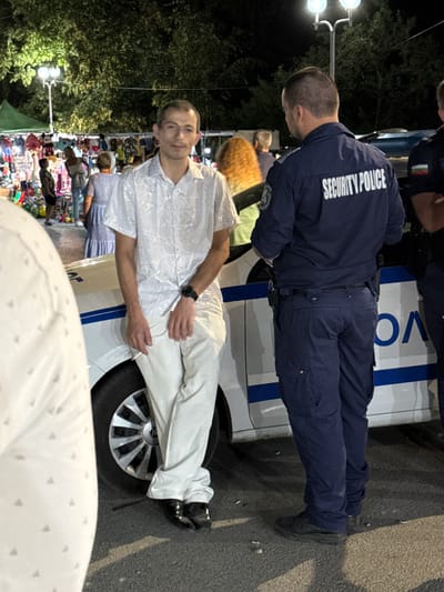 An outdoor scene at night, possibly a market or festival, in Stob, Bulgaria. A man in a white, patterned short-sleeved shirt and light-colored pants leans against a white car with blue stripes and the word "POLICE" visible. He is holding a cigarette. He wears a dark watch. Standing next to him, facing away from the camera, is a person in a navy blue uniform with "SECURITY POLICE" written on the back. The uniform includes a badge on the left sleeve and dark pants. The background shows trees, lighting fixtures, and stalls with merchandise, along with other people milling about.