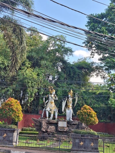 Two statues of what appear to be deities stand on a rock formation in front of dense green foliage. The statue on the left is in a dynamic pose, holding a sword and shield. The statue on the right is also adorned in regal attire. Both statues are white with gold accents. In the foreground, a black metal fence with gold decorative elements encloses a grassy area. Two small trees with red and green foliage are planted on either side of the statues. A red textured wall forms the background behind the statues and foliage. Power lines are strung across the upper portion of the image, against a clear blue sky with scattered clouds. The location is identified as Banjarangkan, Indonesia, with a sign at the base of the rock formation reading "DUMARA."