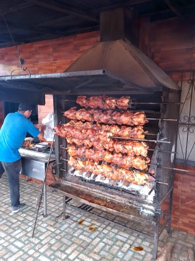 A man in a blue t-shirt and black cap is standing in front of a large rotisserie grill filled with roasting chickens. He is using tongs to remove a chicken. The grill is under a large metal hood, presumably for ventilation. The scene is located outdoors, with tiled flooring and brown tiled walls. The man is working in the city of Villarrica, Paraguay.