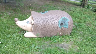 This media file depicts a wooden sculpture of a hedgehog situated in an outdoor grassy area. The sculpture is designed to resemble a log with carved spines and is painted brown. Two stylized green leaves are attached to its back, one of which has a hole in its center. The hedgehog's face is light-colored, with a rounded brown nose and a circular blue eye. It has a visible ear and appears to be resting on two paws. The background consists of green grass, some bare earth, and the base of a tree trunk with foliage visible further back. The lighting suggests it is daytime, and the weather appears mild. There is no visible text in the image. The scene is likely a park or garden in Khmilnyk, Ukraine, given the location context. The sculpture itself is the sole subject, and no activity or people are present.