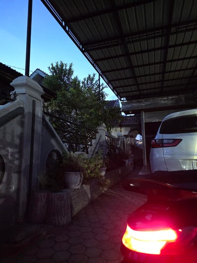 The image depicts an outdoor scene at dusk, likely in a residential area of Bireuen Meunasah Blang, Indonesia. A paved walkway, composed of hexagonal stones, leads towards a house with a tiled roof. To the left, a decorative concrete fence and a gate are visible, partially obscured by lush green foliage from a tree. In the foreground, the red taillight of a motorcycle illuminates the path. To the right, the rear of a silver Honda BR-V SUV is parked under a corrugated metal awning. The sky is a clear, pale blue, transitioning from daylight to evening. The overall impression is that of a quiet, domestic setting at the end of the day.