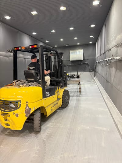 The media file depicts a man operating a yellow forklift inside a large, industrial indoor space. The forklift has "RAEPERS" and "30 LHON" visible on its side. The man is wearing a black t-shirt with text that appears to be "POEDERCONTING KEMPEN" and a black head covering. He is leaning forward, seemingly engaged in a task with the forklift's equipment.
The setting is an industrial interior, possibly a workshop or warehouse, characterized by grey, corrugated walls and a similarly colored ceiling. Numerous square lights are embedded in the wall, illuminating the space. A large, white ventilation unit is visible on the left wall. The floor is concrete and shows tire marks from vehicles. The overall environment appears to be functional and industrial. The lighting suggests an indoor setting, and there are no indicators of the time of day or weather conditions. The scene is focused on the operation of the forklift and the man's involvement with it.