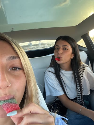 Two young women are depicted inside a passenger vehicle, located in the city of Rosh Haayin, Israel. The image is framed from a close-up, slightly low-angle perspective. In the immediate foreground, the right side of a young woman's face is visible, featuring long blonde hair, a brown eye, and a silver nose ring. Her hand, with light blue nail polish, holds a multi-colored, sugar-coated sour gummy candy (red, pink, and green segments) which she is consuming. Behind her, seated in the vehicle, is a second young woman with long dark hair, wearing a white short-sleeved t-shirt and blue jeans. A black and white striped garment is draped over her left shoulder. She has two red, cherry-shaped gummy candies held between her lips and is looking towards the camera. A silver bracelet is visible on her left wrist, and she wears small hoop earrings. The vehicle's interior features light-colored seating elements and a panoramic sunroof. Through the rear window, multi-lane road traffic and road signs are visible under daylight conditions.