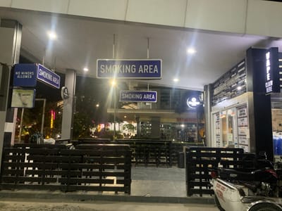 This image depicts an outdoor seating area at night, likely in Angeles, Philippines, with several prominent signs indicating a "SMOKING AREA" and "NO MINORS ALLOWED." The area is furnished with dark wooden benches and stools, suggesting it's a designated space for patrons of nearby establishments.
The scene is illuminated by overhead lights, casting a somewhat dim and moody ambiance. In the background, there are glimpses of shops and businesses. One sign reads "VAPE CLAIM HERE," and another displays "VAP E" vertically, indicating a vape shop or related business. Other text visible includes "MR DIY," "DARTH VAPORS," "SNACKS," "COLD DRINKS," and "COFFEE," further detailing the commercial offerings in the vicinity.
A motorbike, labeled "SKYGO 125," and a white vehicle with "SECURITY AGENCY" written on it are parked in the foreground, hinting at a commercial or mixed-use area. The presence of lights from trees in the background suggests an urban or developed environment. The overall impression is of a commercial strip or plaza at night, with designated smoking zones and various retail outlets. There are no people clearly visible in the image, though a silhouette of people can be seen in the background seating area of a building.