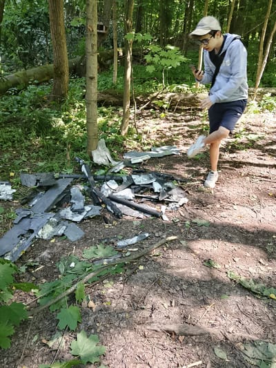In Motyakovo, Russia, a young person wearing a light grey hoodie, dark shorts, glasses, and a beige baseball cap is standing in a wooded area. They are looking down at a smartphone in their left hand and appear to be interacting with it while lifting their right leg, with their foot slightly extended toward a pile of debris. The debris consists of dark, irregularly shaped fragments, possibly roofing materials, scattered on the ground among leaves and twigs. The environment is a forest floor with trees and dense undergrowth, illuminated by dappled sunlight. A fallen, moss-covered log is visible in the background, along with a small wooden birdhouse attached to a tree.
